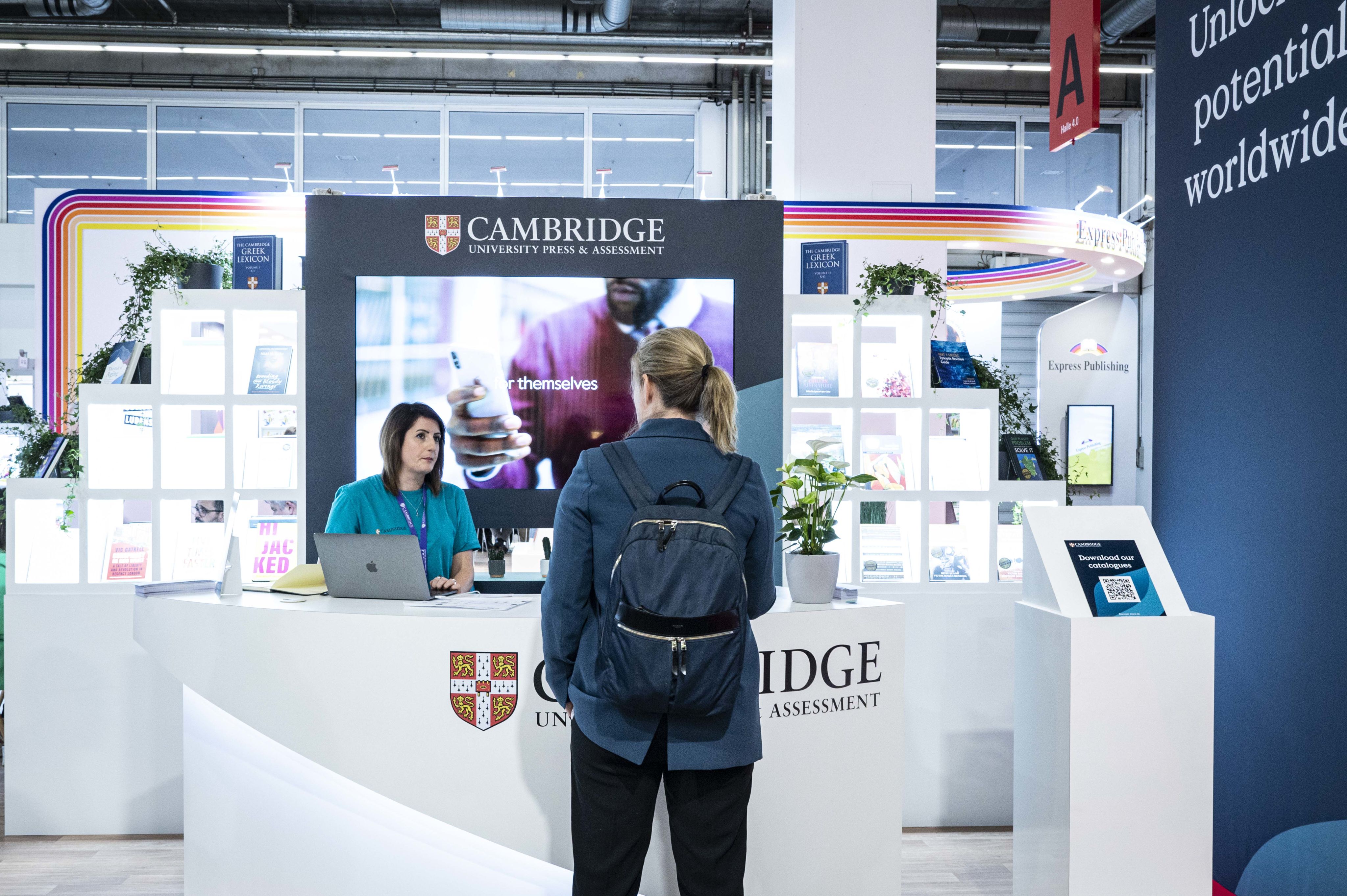 a woman behind a desk at the Cambridge booth at a book fair speaks to another woman visiting the desk