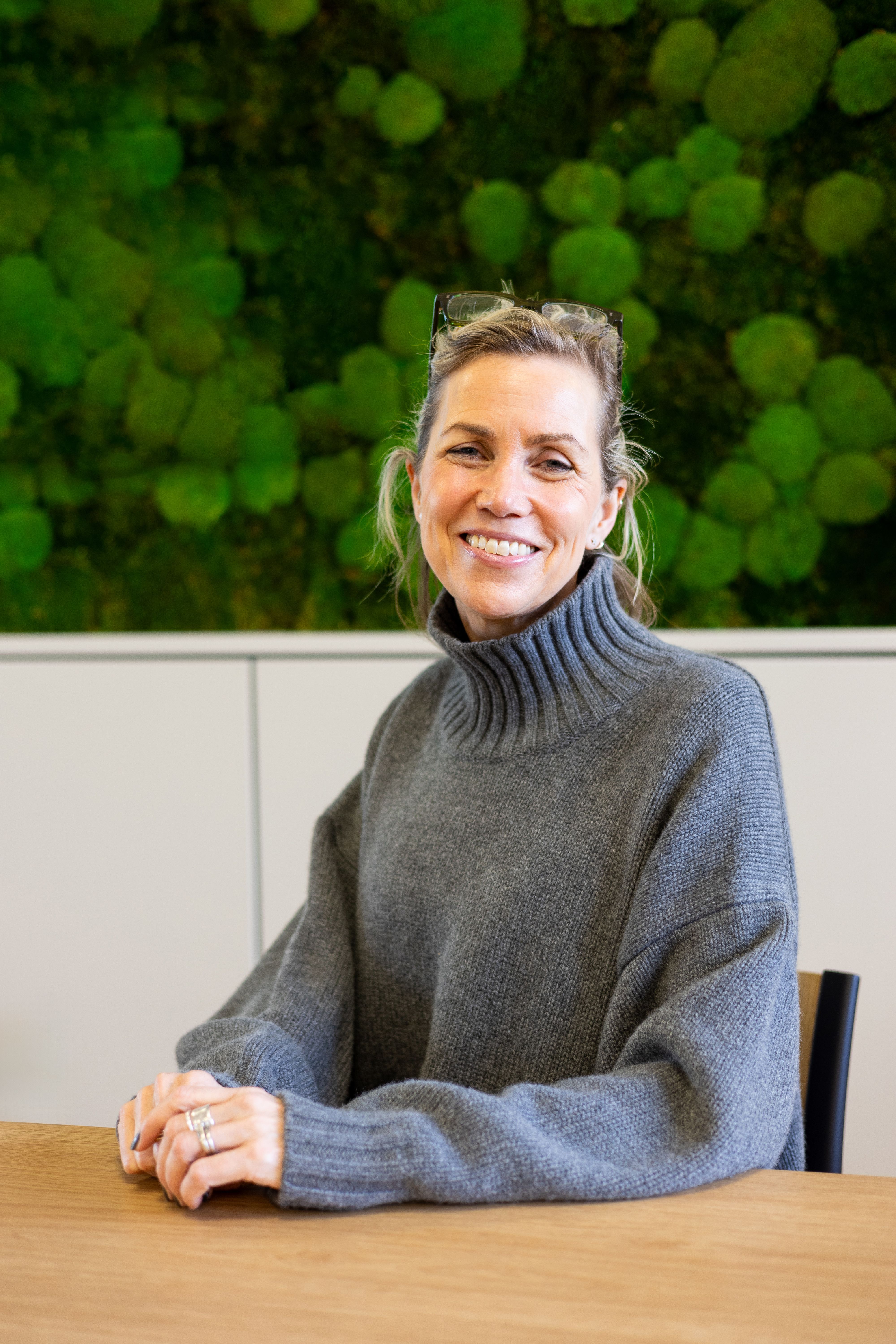 Julie Dennis sat at a table smiling at the camera with a green background behind her