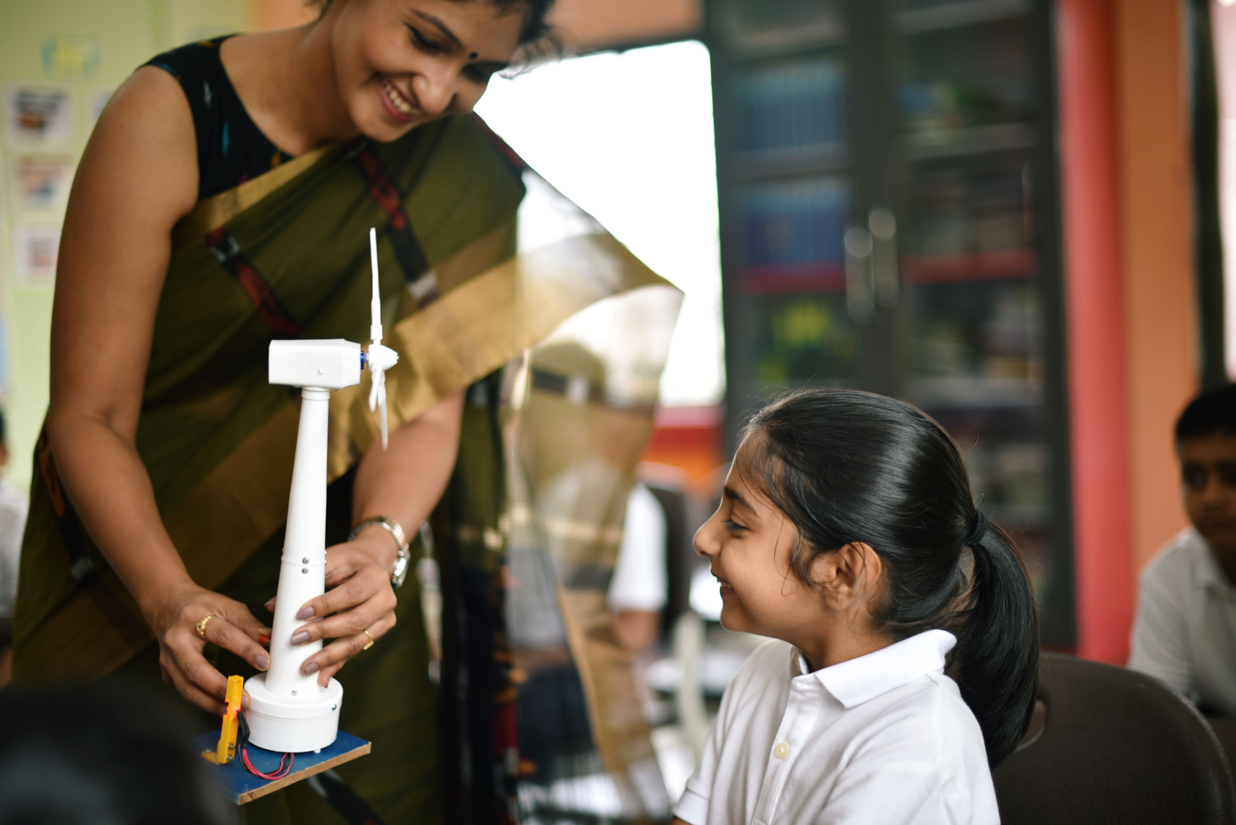 teacher wearing a sari smiles while holding a model of a wind turbine in front of a smiling student in a white school uniform